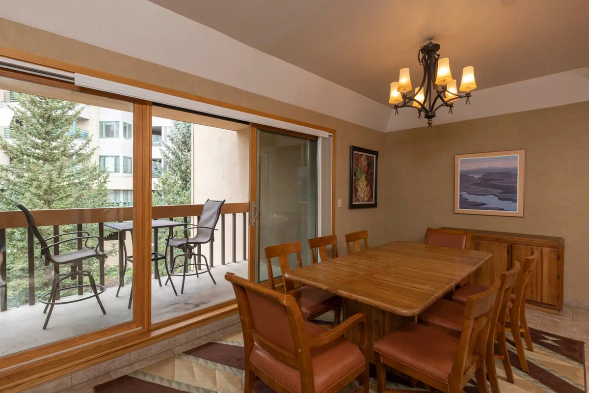 This image features a spacious dining area centered around a wooden table beneath a stylish chandelier. The large balcony window reveals scenic mountain views, and the wall art and décor add warmth. The setting is perfect for family meals or celebratory gatherings.