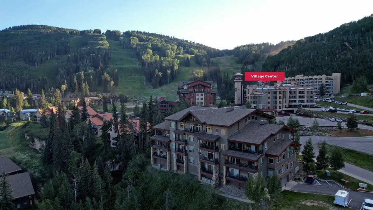 A wide exterior view of the Village Center set against a breathtaking mountain backdrop, surrounded by trees and neighboring residential buildings. The image communicates the resort's central, family-friendly location and the stunning natural environment that frames every stay.