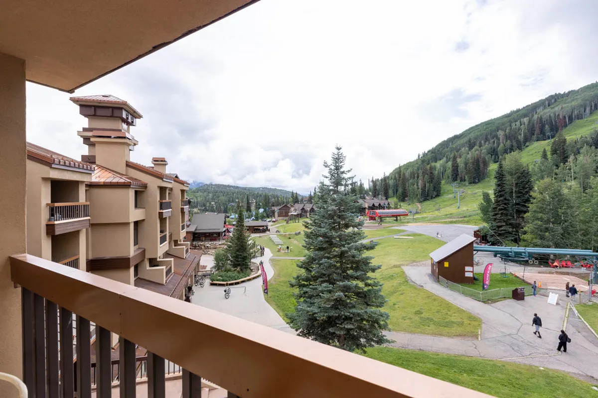 This image captures a sweeping view from a balcony, with a ski lift visible in the distance, walking paths winding through lush greenery, and adjacent buildings that hint at the lively atmosphere of the mountain village. The scene conveys both the thrill of winter sports and the relaxed energy of summer activities, making it perfect for showcasing year-round adventure.
