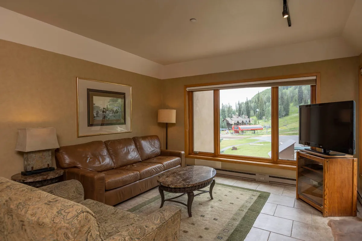 A welcoming living area featuring a leather sofa positioned to enjoy a sweeping panoramic view through expansive windows, with a coffee table in the center. The scene is cozy and captures a sense of relaxation and togetherness, with natural light pouring in and the mountains visible beyond.