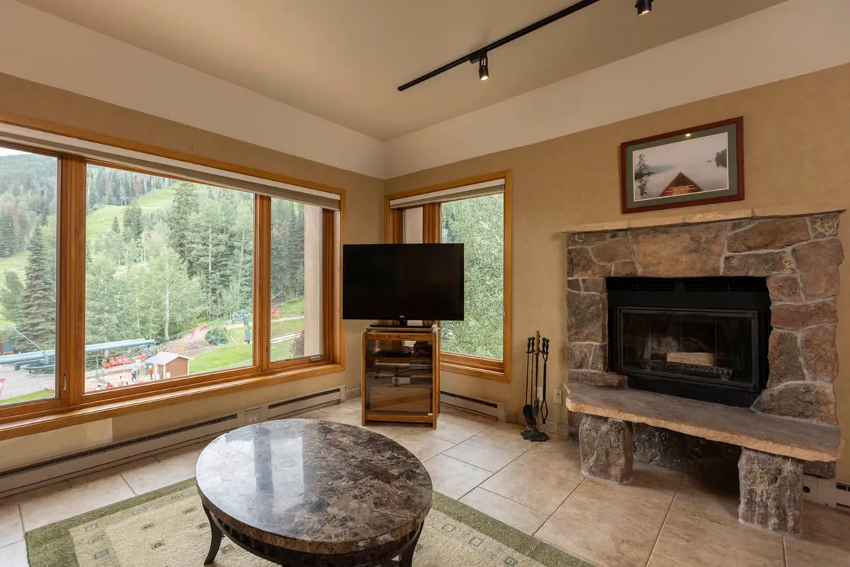 A broad view of the living space, centered around a striking stone fireplace and large windows that flood the room with light and frame the surrounding mountain landscape. The setup encourages both relaxation and socializing, featuring comfortable seating and a television for leisure.