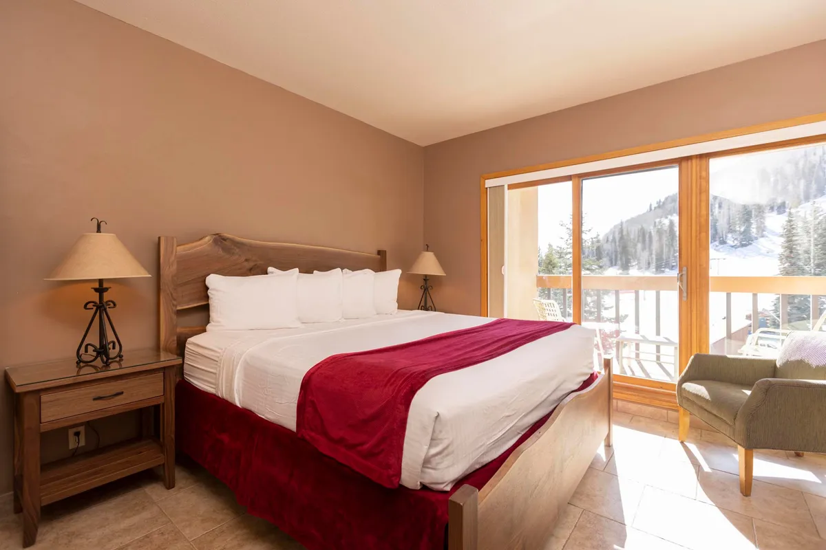 A tranquil bedroom featuring a large bed with white linens and a red accent, flanked by wooden side tables with lamps. A large window offers breathtaking mountain views, allowing plenty of natural light to fill the room and enhancing the sense of comfort and serenity.