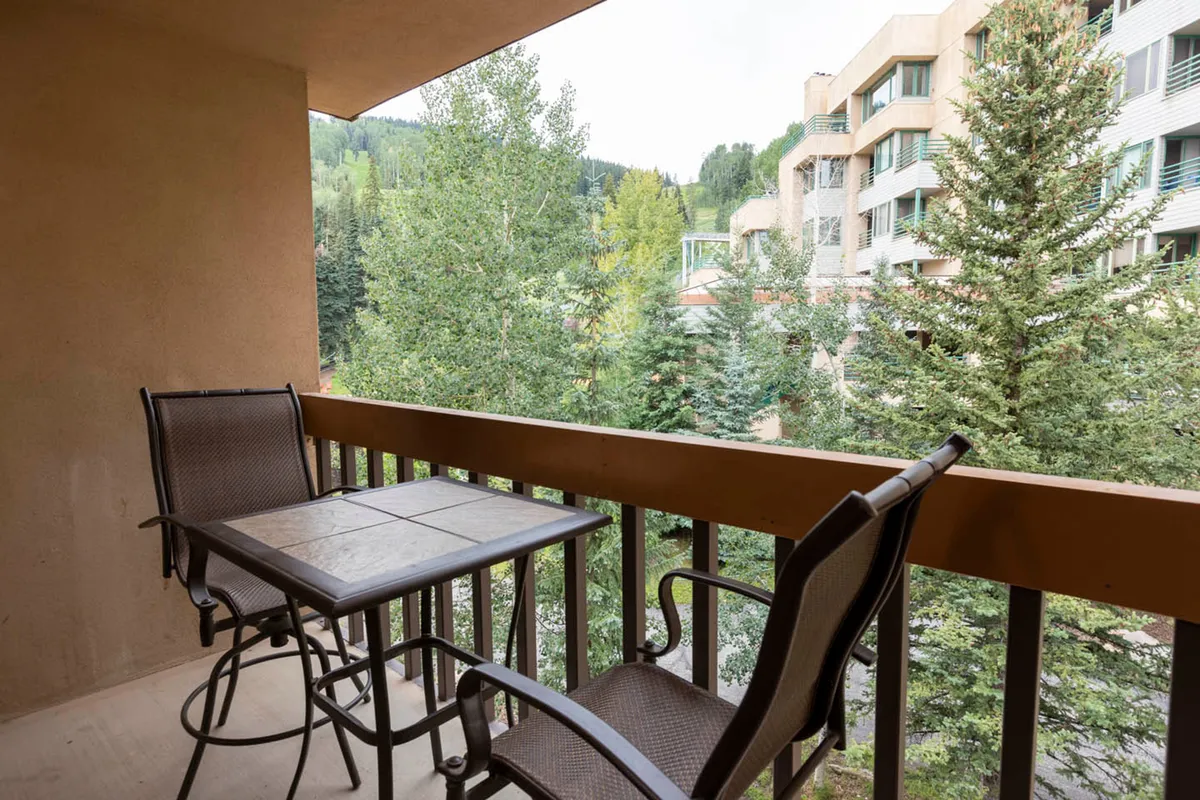 An exterior view featuring outdoor balcony seating surrounded by lush trees and framed by a distant mountain view. The scene is serene, suggesting both a sense of seclusion and connection to nature. The space is ideal for morning coffee, reading, or simply soaking in the mountain atmosphere.