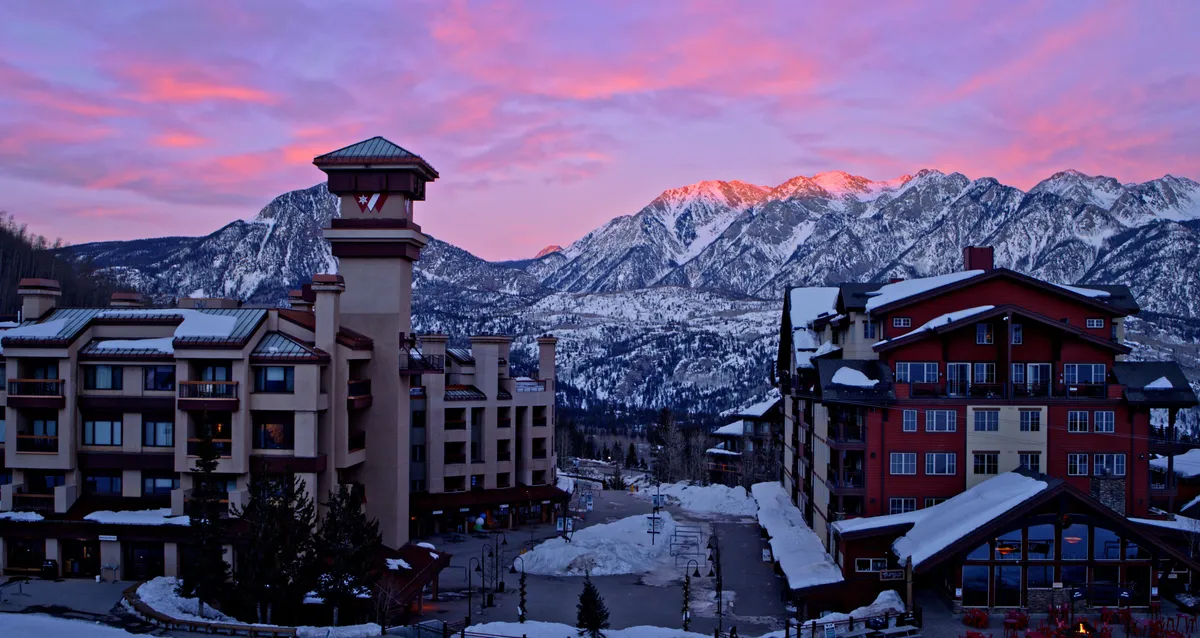 A wide exterior shot displaying the majestic mountains, property buildings, and a glowing sunset sky. The scene conveys the proximity to outdoor adventures like skiing and snowboarding, while also suggesting the warmth and coziness awaiting guests after a day out. The overall mood is serene and welcoming.