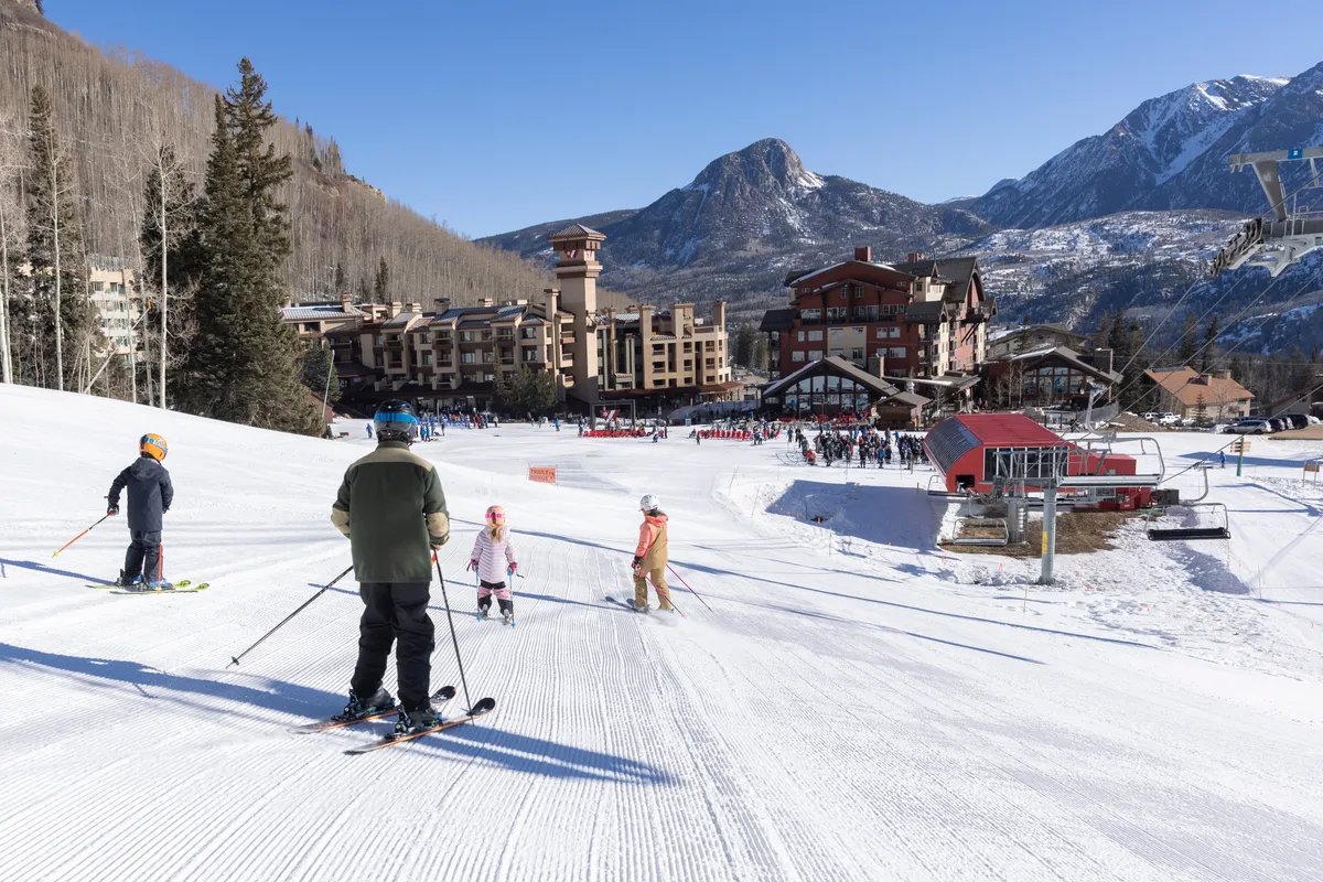 This image captures skiers in motion on a snow-covered slope, with the resort building positioned at the base and rugged mountains rising in the background. The lively winter scene conveys both the direct slope access and active mountain lifestyle promoted in the section.