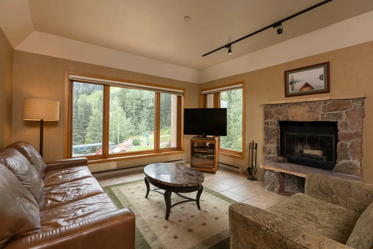 The image showcases a cozy living area with a stunning stone fireplace, plush seating, and expansive windows framing breathtaking mountain and ski slope views. The room is bathed in natural light, inviting guests to relax, watch the sunset, and enjoy time together in a warm, inviting atmosphere.