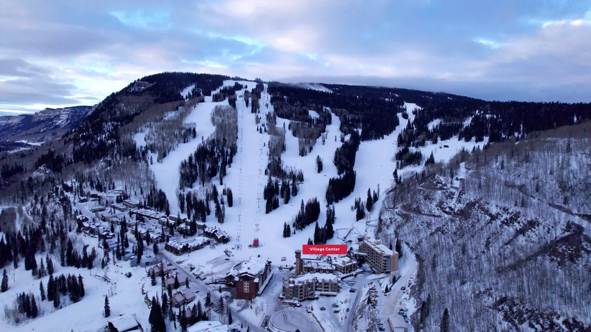 A sweeping exterior view of ski slopes descending towards a charming village center, encircled by snow-covered trees and majestic mountains. The inviting winter ambiance hints at après-ski gatherings, exploration, and mountain relaxation.
