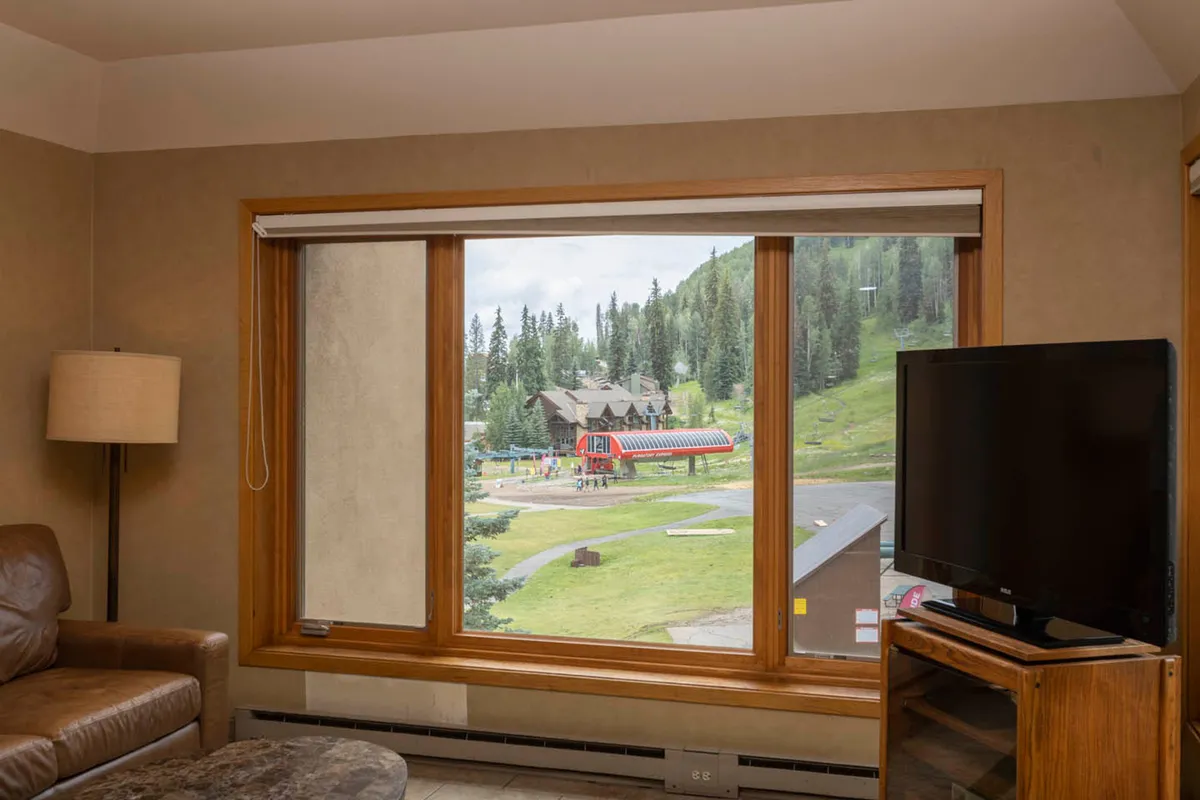 A comfortable interior lounge area with a large window framing the mountains, a plush sofa, and a television, creating a welcoming spot to relax after outdoor adventures. Sunlight highlights the interior's inviting textures and the ever-present wilderness beyond.