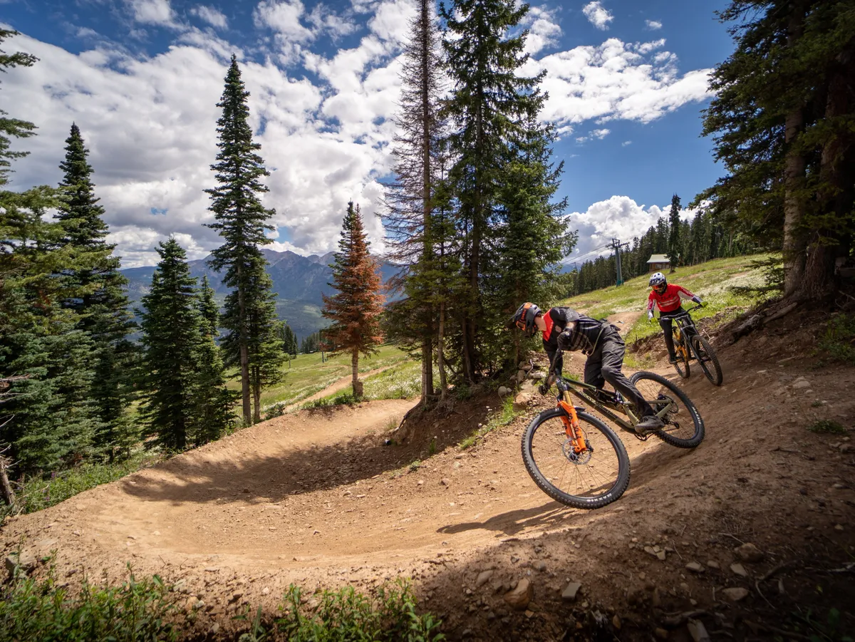 An action-filled shot of mountain bikers navigating a winding trail through tall trees, with the majestic mountains forming the backdrop. The energy and vibrancy of the scene evoke the spirit of outdoor adventure available during warmer months.